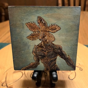 May include: A wooden block featuring a detailed pyrography image of a Demogorgon from the popular science fiction horror series. The artwork is in shades of brown and black against a teal background.