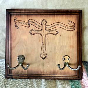 May include: Wooden wall-mounted coat rack with a dark brown frame. The light-colored wood features a cross and banner with text from Luke 10:5. Two silver hooks are attached below the cross for hanging items.