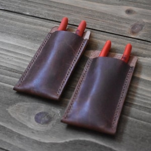 May include: Two brown leather pouches with red tool tips sticking out of the top. The pouches are stitched with a decorative stitch.