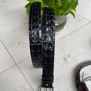 Op de afbeelding: Een zwarte leren riem met een textuur die op krokodillenleer lijkt. De riem heeft een zilverkleurige rechthoekige gesp. De riem wordt getoond tegen een neutrale achtergrond, wat het ontwerp en de afwerking benadrukt.