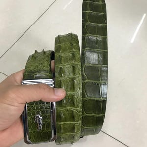 May include: A green alligator leather belt with a silver buckle. The belt is made of genuine alligator skin and has a unique, textured pattern.