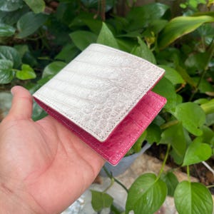 Peut inclure: Un portefeuille à deux volets avec un extérieur blanc texturé et un intérieur rose. Le portefeuille est ouvert, révélant les couleurs et les textures contrastées des matériaux. Le design suggère un accent sur le luxe et le savoir-faire.
