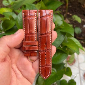 Könnte beinhalten: Zwei braune Lederarmbänder mit weißen Nähten. Die Armbänder sind aus Alligatorleder und haben ein klassisches Design.