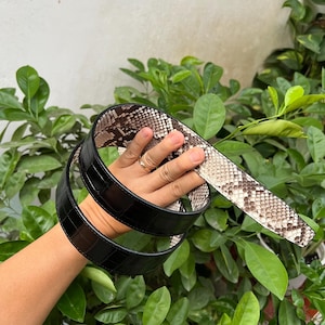 以下が含まれることがあります： ヘビ柄の黒と茶色の革ベルト。ベルトは半分に折り畳まれ、手で持たれています。