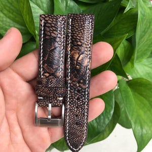 May include: A brown leather watch strap with white stitching detail. The strap has a textured pattern resembling scales.