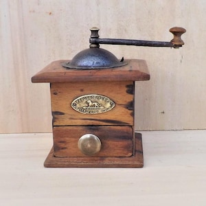 May include: A vintage wooden coffee grinder with a metal crank and a small drawer. The grinder has a brass knob and a decorative metal plate on the front. The wood has a warm, natural tone.