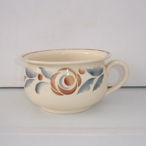 May include: A white ceramic chamber pot with a blue and brown floral design. The pot has a handle on the side.