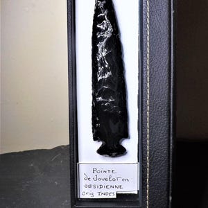 May include: A glossy black obsidian arrowhead displayed in a black box with a white interior. The arrowhead is sharply pointed and has a textured surface. A label in the box reads "Pointe de Javelot en Obsidienne Orig. INDES."
