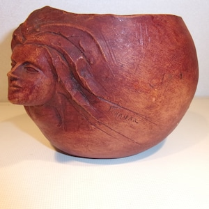 Könnte beinhalten: Ein brauner Keramiktopf mit einem geschnitzten Frauenkopf an der Seite. Der Topf ist mit "Thanar" signiert.
