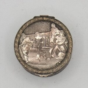 May include: A silver compact with a detailed engraved design featuring a scene of people in a room. The compact has a circular shape and a raised rim.