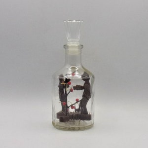 Puede incluir: Una botella de vidrio transparente con un tapón decorativo y una escena en el interior que representa dos figuras y un pequeño árbol con bayas rojas.