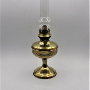 Puede incluir: Una l&aacute;mpara de aceite de lat&oacute;n con una chimenea de vidrio transparente. La l&aacute;mpara tiene una base redonda y un dise&ntilde;o decorativo en la parte superior.