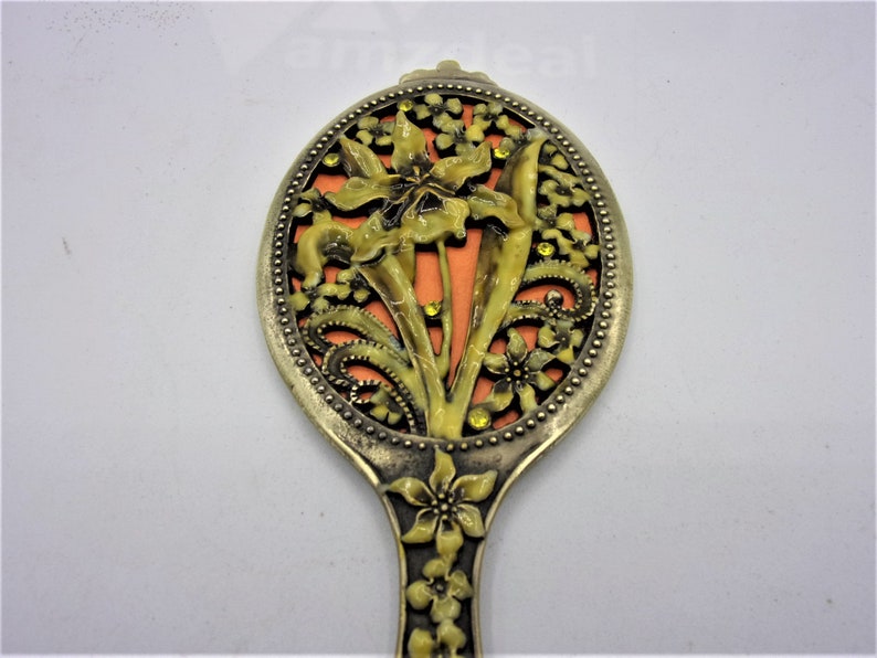 May include: A vintage hand mirror with a floral design. The mirror is made of metal and has a gold finish. The floral design is in shades of yellow and orange and features a large flower in the center.