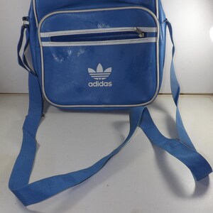 May include: A blue Adidas shoulder bag with a white logo and white trim. The bag has a zippered pocket on the front.