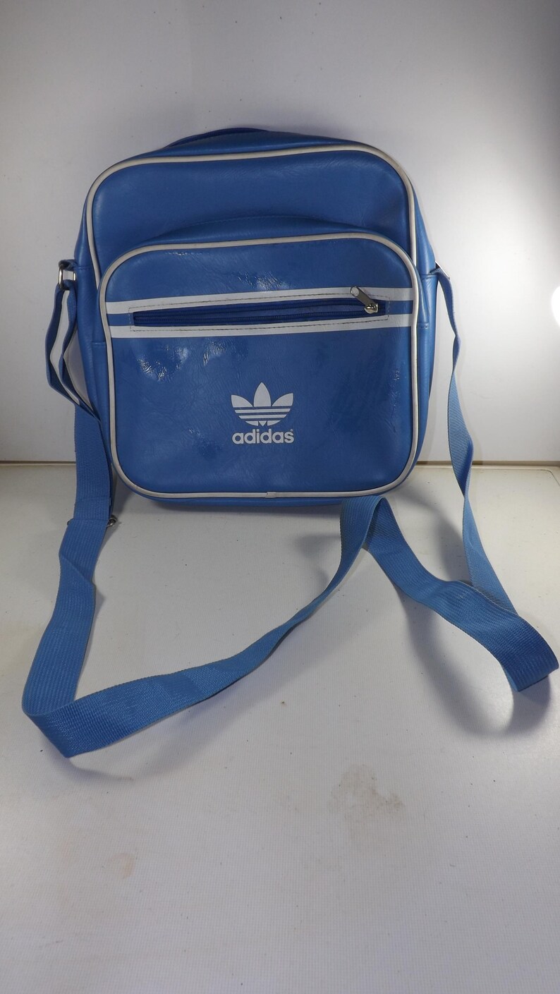 May include: A blue Adidas shoulder bag with a white logo and white trim. The bag has a zippered pocket on the front.