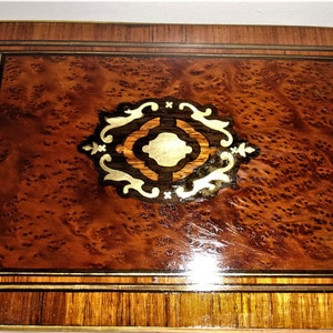 May include: A wooden box with a marquetry design on the top. The design features a central circle with a geometric pattern surrounded by floral motifs. The box is made of dark wood with a reddish brown hue.