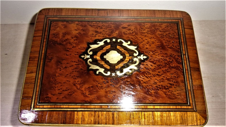 May include: A wooden box with a marquetry design on the top. The design features a central circle with a geometric pattern surrounded by floral motifs. The box is made of dark wood with a reddish brown hue.