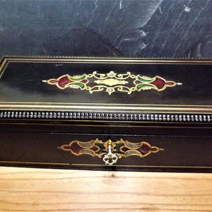May include: A rectangular black box with ornate gold, red, and green decorative accents. The box has a hinged lid and a decorative keyhole. The box is sitting on a wooden surface.