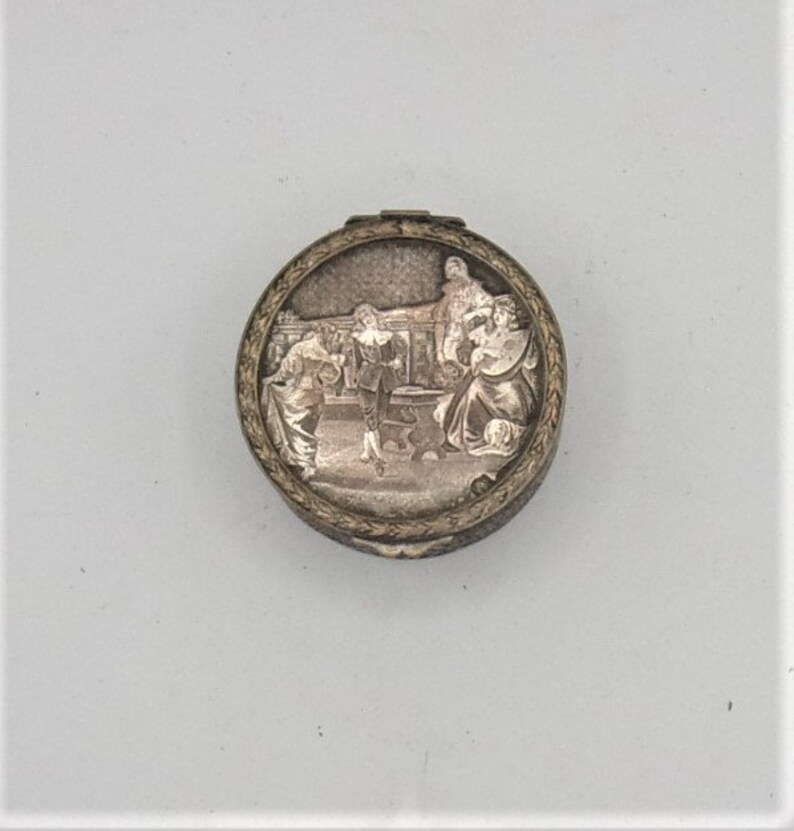 May include: A silver compact with a detailed engraved design featuring a scene of people in a room. The compact has a circular shape and a raised rim.