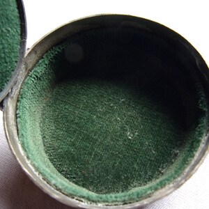 May include: A small, round, silver-colored box with a green velvet lining. The box is open and the lining is visible.