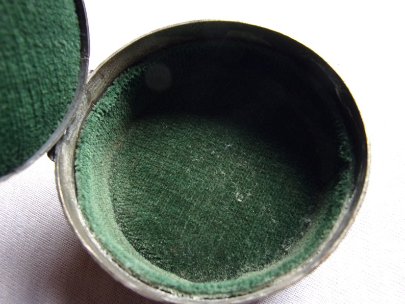 May include: A small, round, silver-colored box with a green velvet lining. The box is open and the lining is visible.