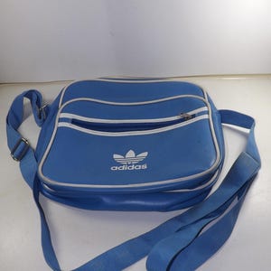 May include: A blue Adidas shoulder bag with a white stripe and the Adidas logo. The bag has a zippered pocket on the front.