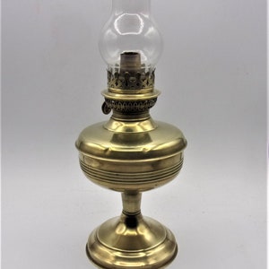 Puede incluir: Una l&aacute;mpara de aceite de lat&oacute;n con una chimenea de vidrio transparente. La l&aacute;mpara tiene una base redonda y un dise&ntilde;o decorativo alrededor de la parte superior.