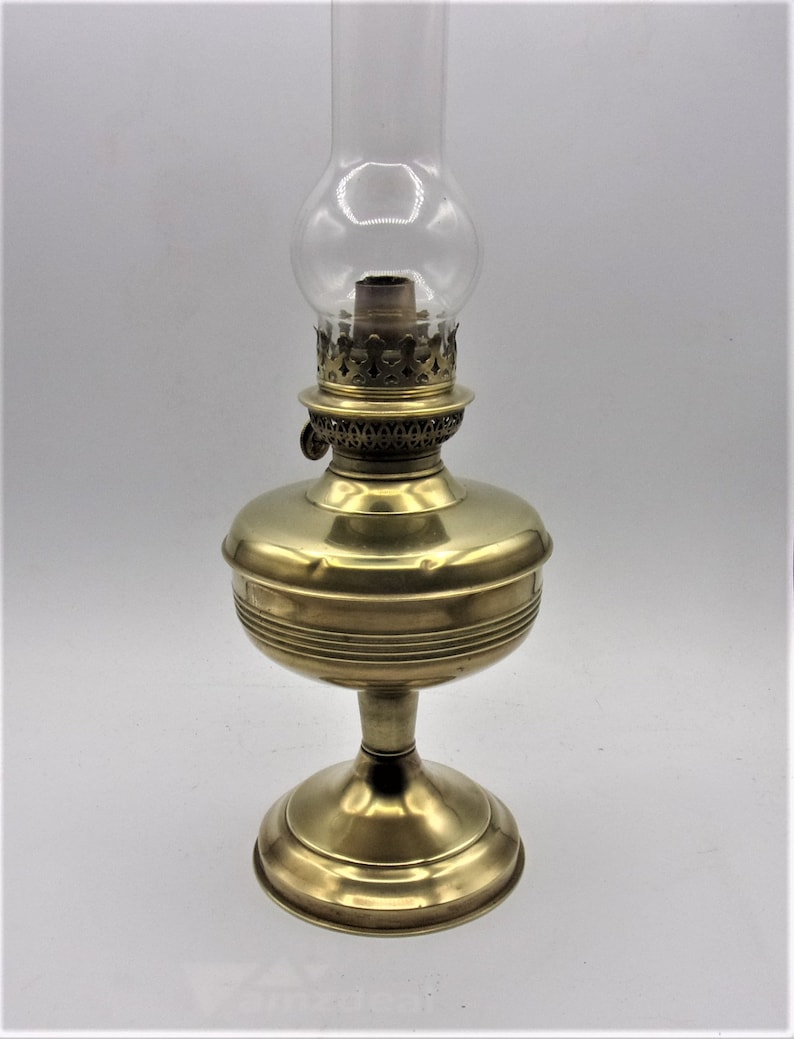 Puede incluir: Una l&aacute;mpara de aceite de lat&oacute;n con una chimenea de vidrio transparente. La l&aacute;mpara tiene una base redonda y un dise&ntilde;o decorativo alrededor de la parte superior.