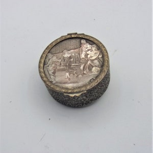 May include: A round, silver-colored trinket box with an ornate design. The lid features a raised image of a group of people in a scene.