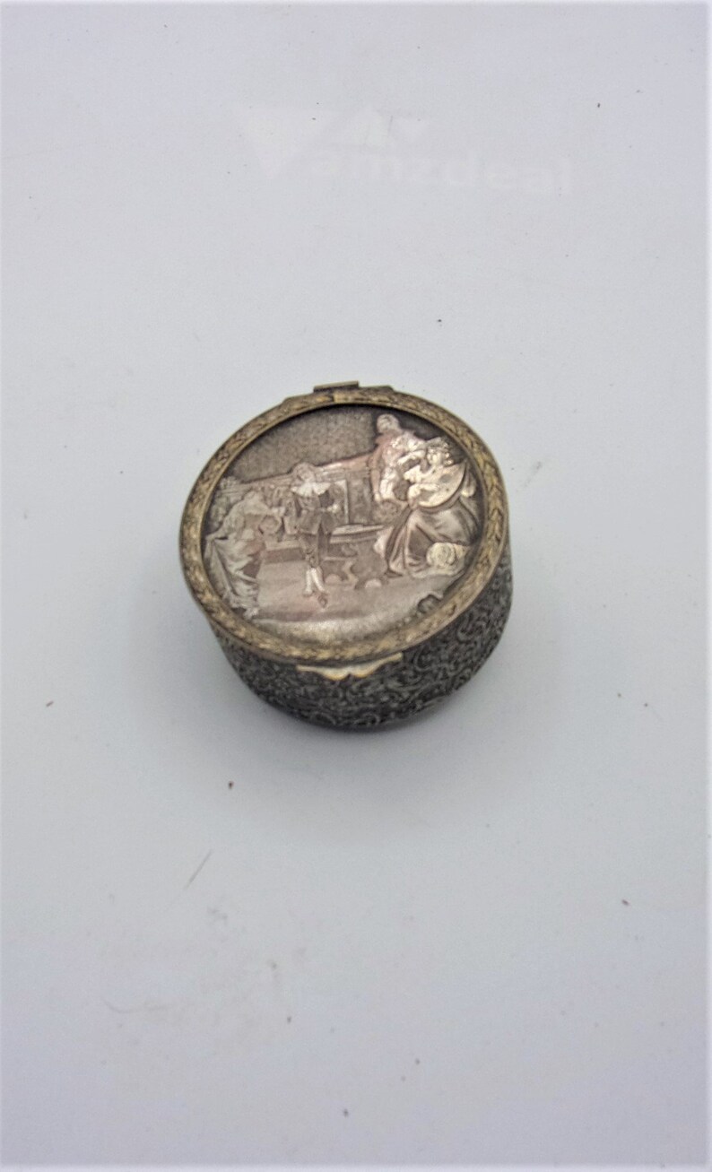 May include: A round, silver-colored trinket box with an ornate design. The lid features a raised image of a group of people in a scene.