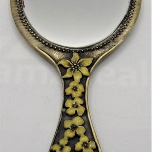 May include: A vintage hand mirror with a round, silver-toned frame and a floral design on the handle. The handle is decorated with yellow flowers.