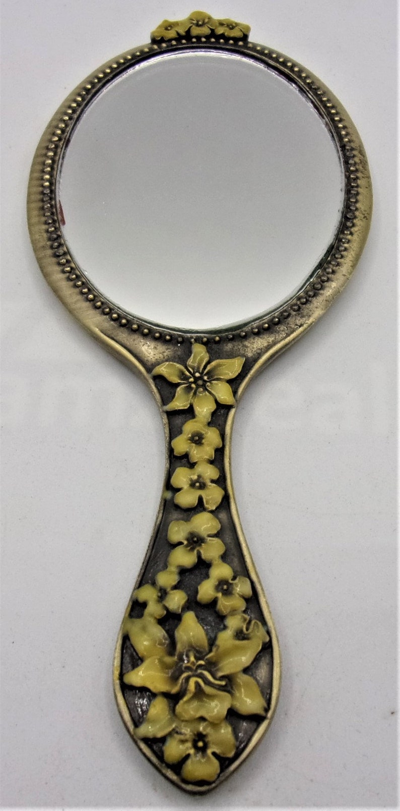 May include: A vintage hand mirror with a round, silver-toned frame and a floral design on the handle. The handle is decorated with yellow flowers.