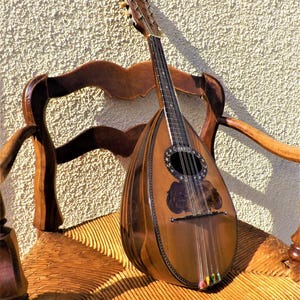 Può includere: Un mandolino vintage è appoggiato su una sedia di legno. Il mandolino ha una finitura marrone scuro con una buca decorativa e corde. La sedia è in legno con una seduta intrecciata, che mostra un'estetica rustica.