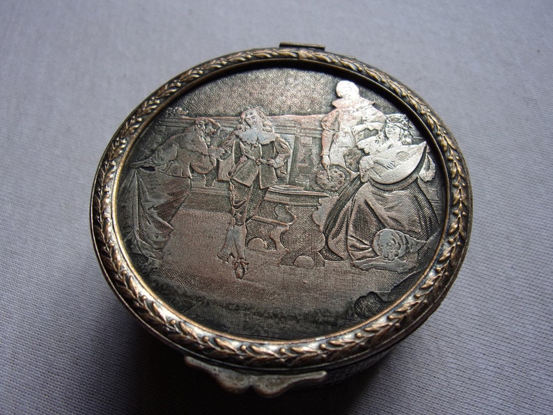 May include: A silver and gold circular compact with an embossed design of four people in period clothing. The compact has a decorative border.