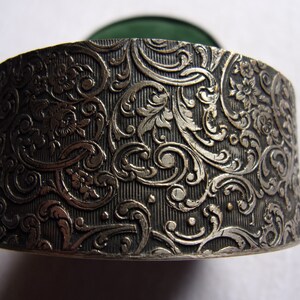 May include: A silver round box with an intricate floral design. The box has a green velvet interior.