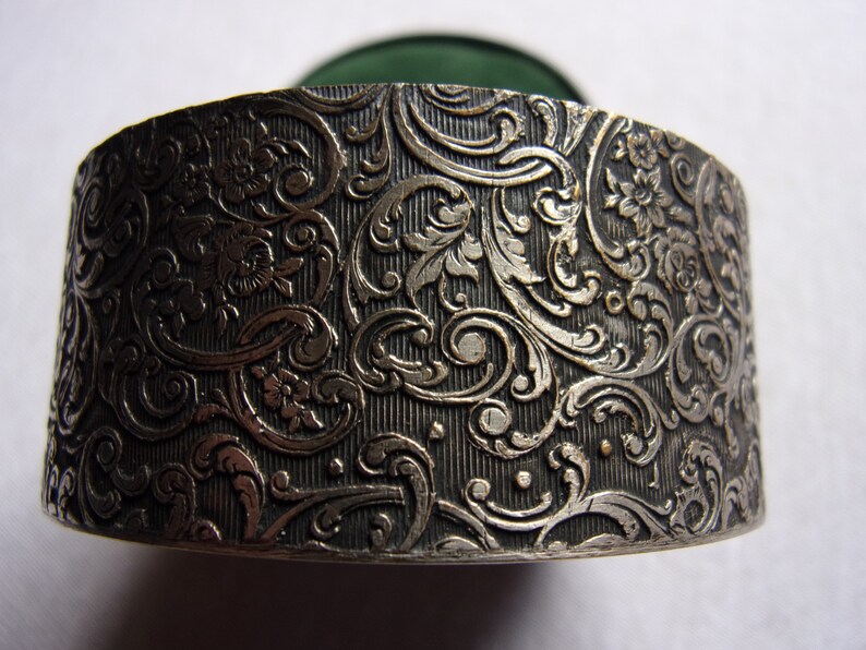 May include: A silver round box with an intricate floral design. The box has a green velvet interior.