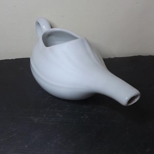 May include: A white ceramic neti pot with a handle and a spout. The pot is designed with a rounded body and a long, slender spout. It is placed on a dark surface.