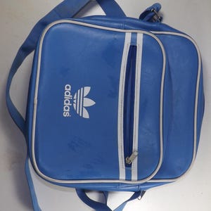 May include: A blue Adidas shoulder bag with a white zipper and white Adidas logo. The bag has a white stripe down the side.