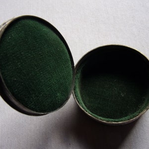 May include: A round, silver-colored trinket box with a hinged lid. The inside of the box is lined with dark green velvet.