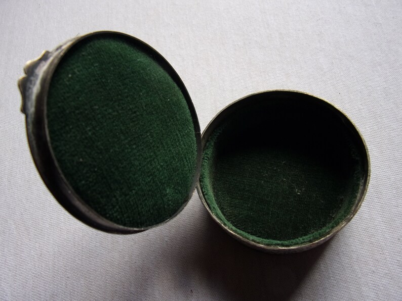 May include: A round, silver-colored trinket box with a hinged lid. The inside of the box is lined with dark green velvet.