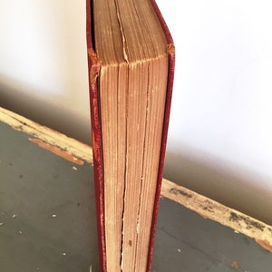 Może przedstawiać: Antyczna książka w twardej oprawie z czerwoną okładką i postarzanymi stronami. Książka stoi pionowo na drewnianej powierzchni.