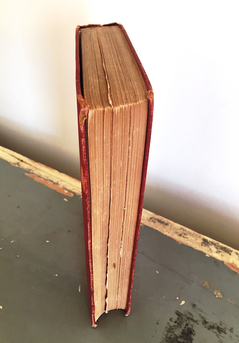 Może przedstawiać: Antyczna książka w twardej oprawie z czerwoną okładką i postarzanymi stronami. Książka stoi pionowo na drewnianej powierzchni.