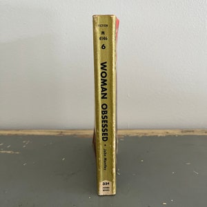 May include: A gold paperback book with the title "Woman Obsessed" by John Manley. The book is part of the Perma Books series and has a fiction category label.