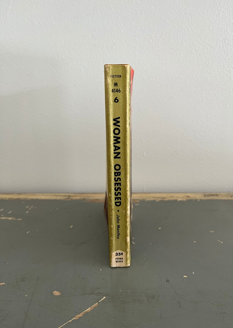 May include: A gold paperback book with the title "Woman Obsessed" by John Manley. The book is part of the Perma Books series and has a fiction category label.