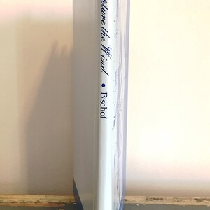 May include: A white book with a blue spine and red top. The title of the book is "Capture the Wind" by Bischof. The book is on a wooden surface.