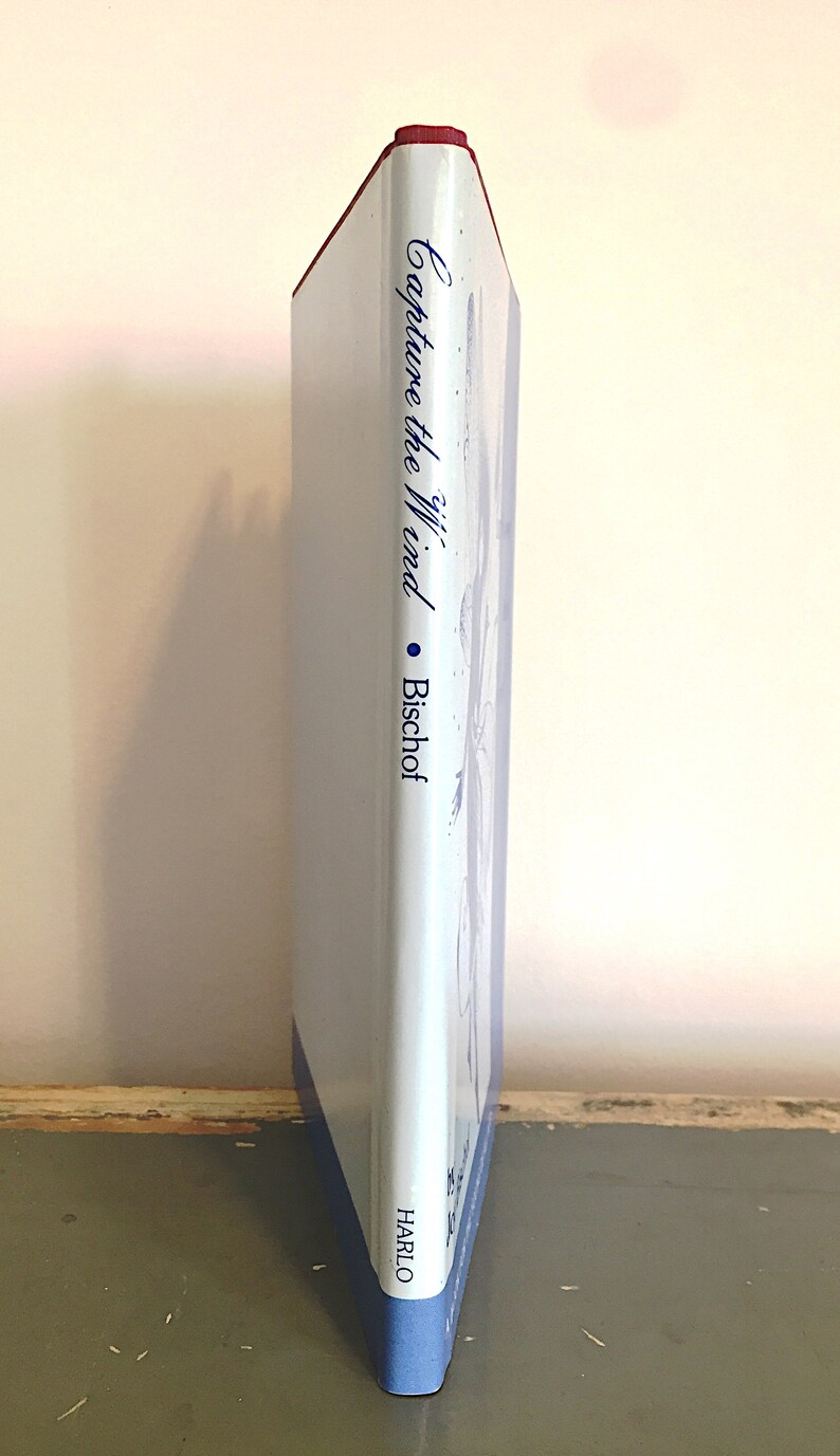 May include: A white book with a blue spine and red top. The title of the book is "Capture the Wind" by Bischof. The book is on a wooden surface.