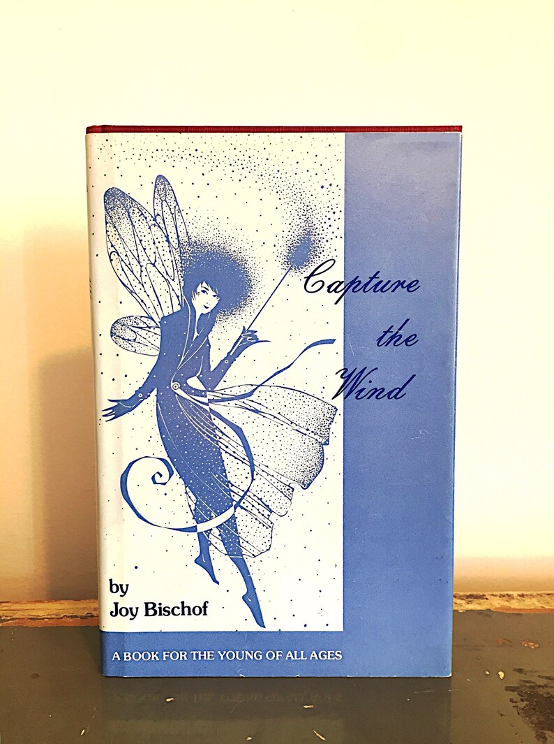 May include: A book cover with a blue and white illustration of a fairy with wings. The text on the cover reads "Capture the Wind" by Joy Bischof. The book is described as "A book for the young of all ages."
