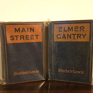Puede incluir: Dos libros antiguos, "Main Street" y "Elmer Gantry" de Sinclair Lewis, están envueltos en plástico. Las portadas son de color azul marino con etiquetas naranjas que llevan los títulos en texto blanco en negrita. El nombre del autor está impreso en naranja.