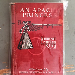 Może przedstawiać: Czerwona książka zatytułowana "An Apache Princess" autorstwa generała Charlesa Kinga. Okładka książki przedstawia ilustrację indiańskiego nakrycia głowy i fajki. Książkę ilustrowali Frederic Remington i E.W. Deming.