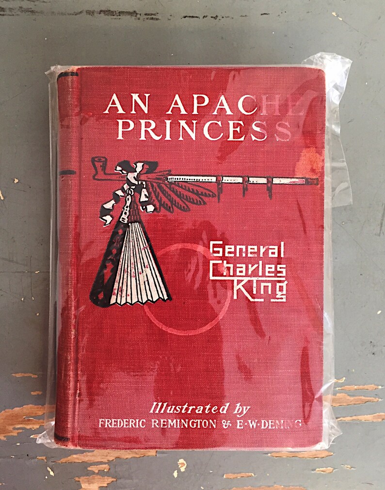 Może przedstawiać: Czerwona książka zatytułowana "An Apache Princess" autorstwa generała Charlesa Kinga. Okładka książki przedstawia ilustrację indiańskiego nakrycia głowy i fajki. Książkę ilustrowali Frederic Remington i E.W. Deming.